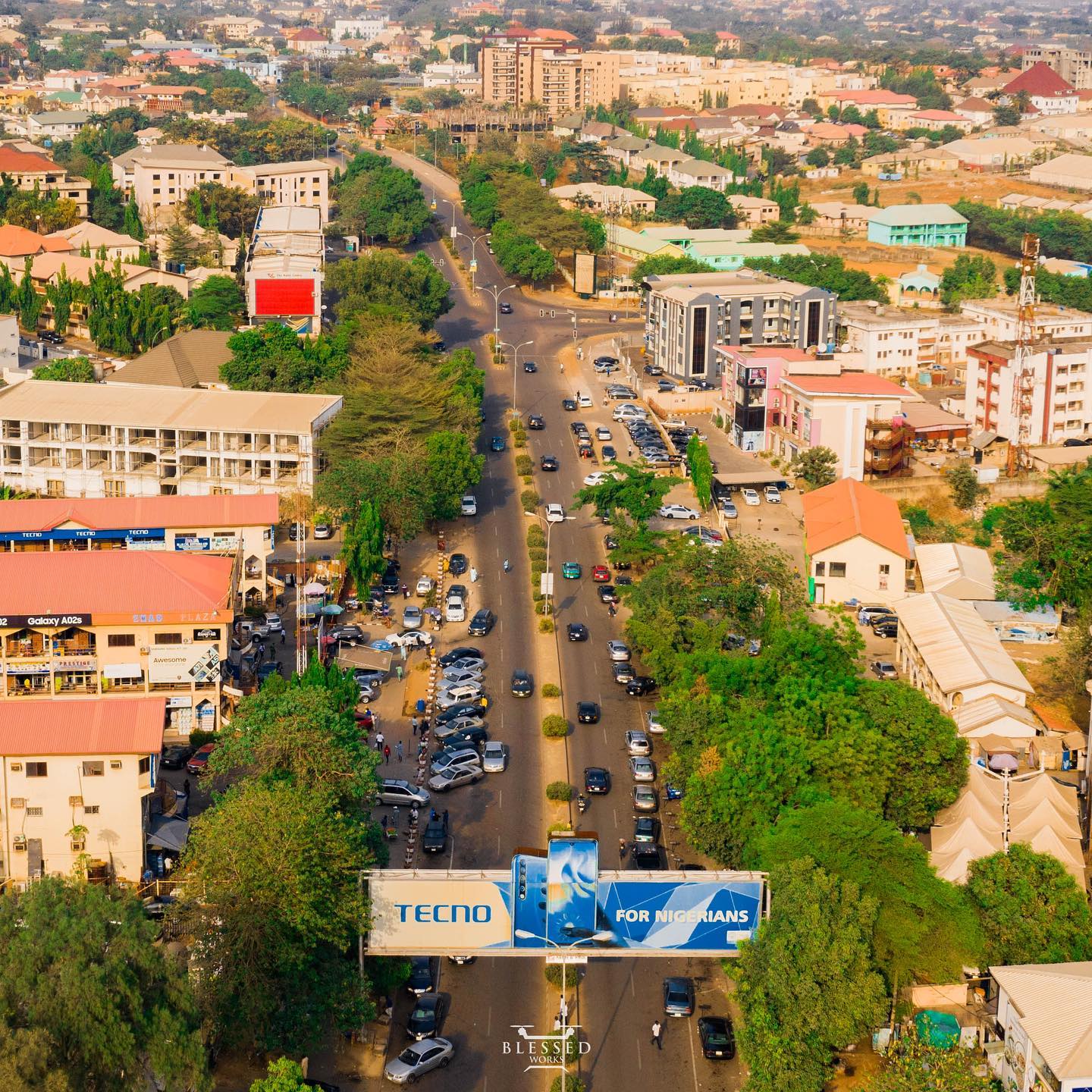 Aerial drone view of a busy commercial street in Wuse, Abuja, Nigeria, lined with trees, multi-storey buildings, parked cars, and a large Tecno billboard reading "For Nigerians" in the foreground.