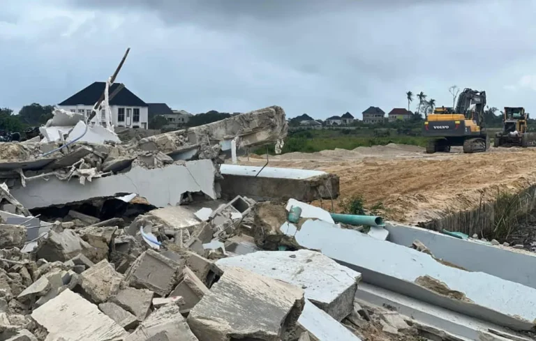 Demolished buildings at WinHomes 2 estate showing rubble, broken concrete, and exposed pipes in the foreground, with a Volvo excavator clearing sandy ground and residential houses visible in the background.