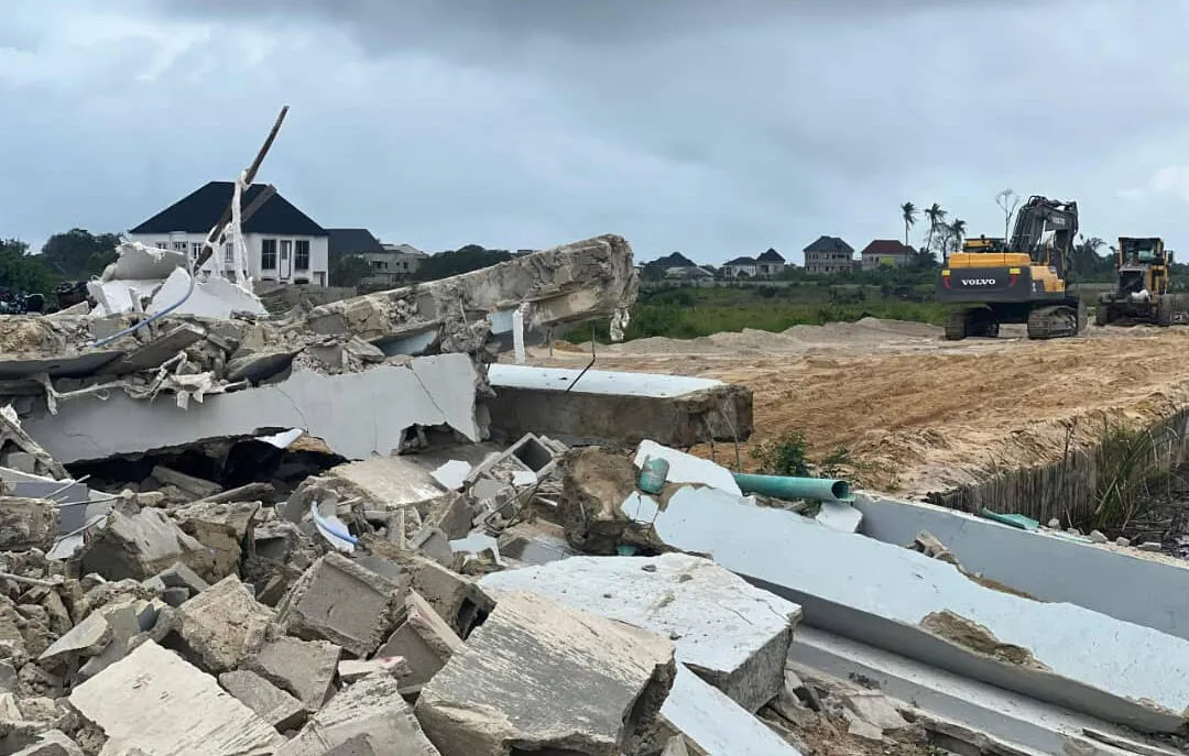 Demolished buildings at WinHomes 2 estate showing rubble, broken concrete, and exposed pipes in the foreground, with a Volvo excavator clearing sandy ground and residential houses visible in the background.