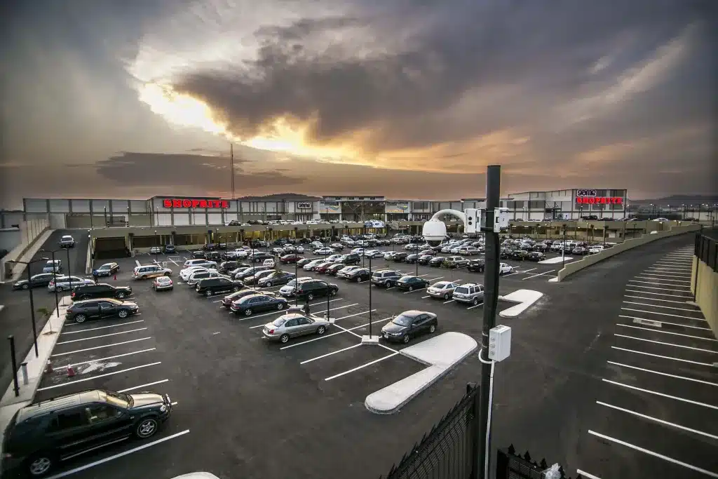 A wide view of Jabi Lake Mall in Abuja showing a large parking lot filled with vehicles, retail stores, and shoppers under a dramatic evening sky.
