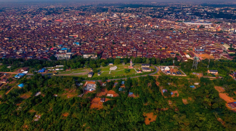 Aerial view of a modern residential estate in Ibadan, Nigeria, featuring well-planned streets, landscaped green spaces, detached homes with solar panels, and surrounding hills.