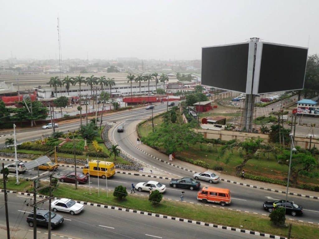 Aerial view of a major road intersection in Ikeja GRA, Lagos, showing moving traffic, landscaped medians, commercial buildings, and surrounding urban infrastructure.