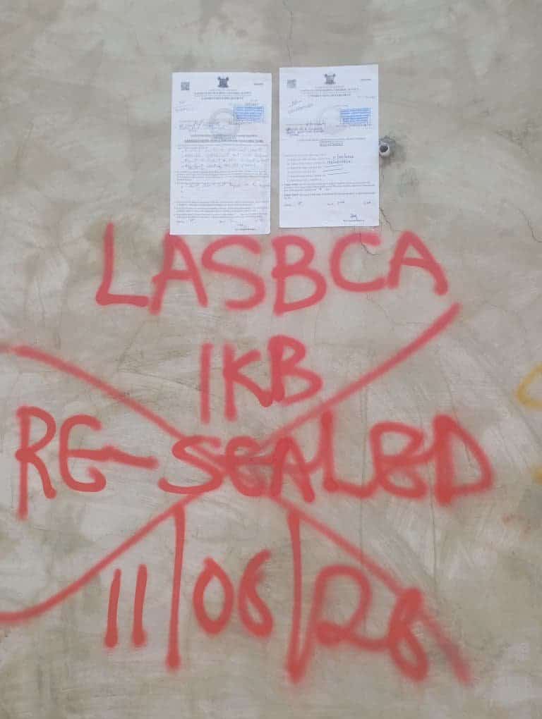 A building wall spray-painted in red with the text "LASBCA 1KB RE-SEALED 11/06/26" and a large red X, with two official government enforcement notices pinned above it.