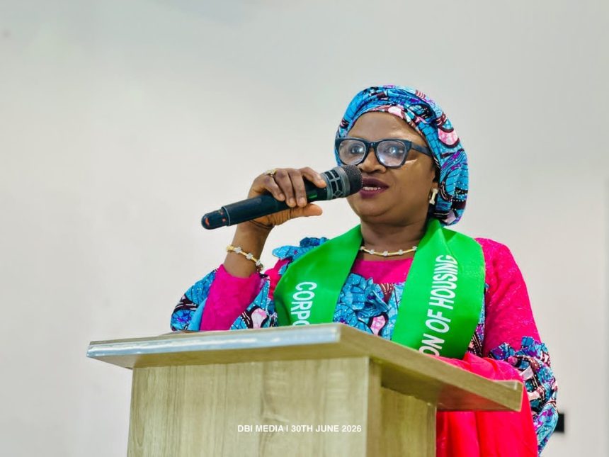 "Woman in colourful traditional attire and a sash speaking into a microphone at a podium during a housing-focused event."